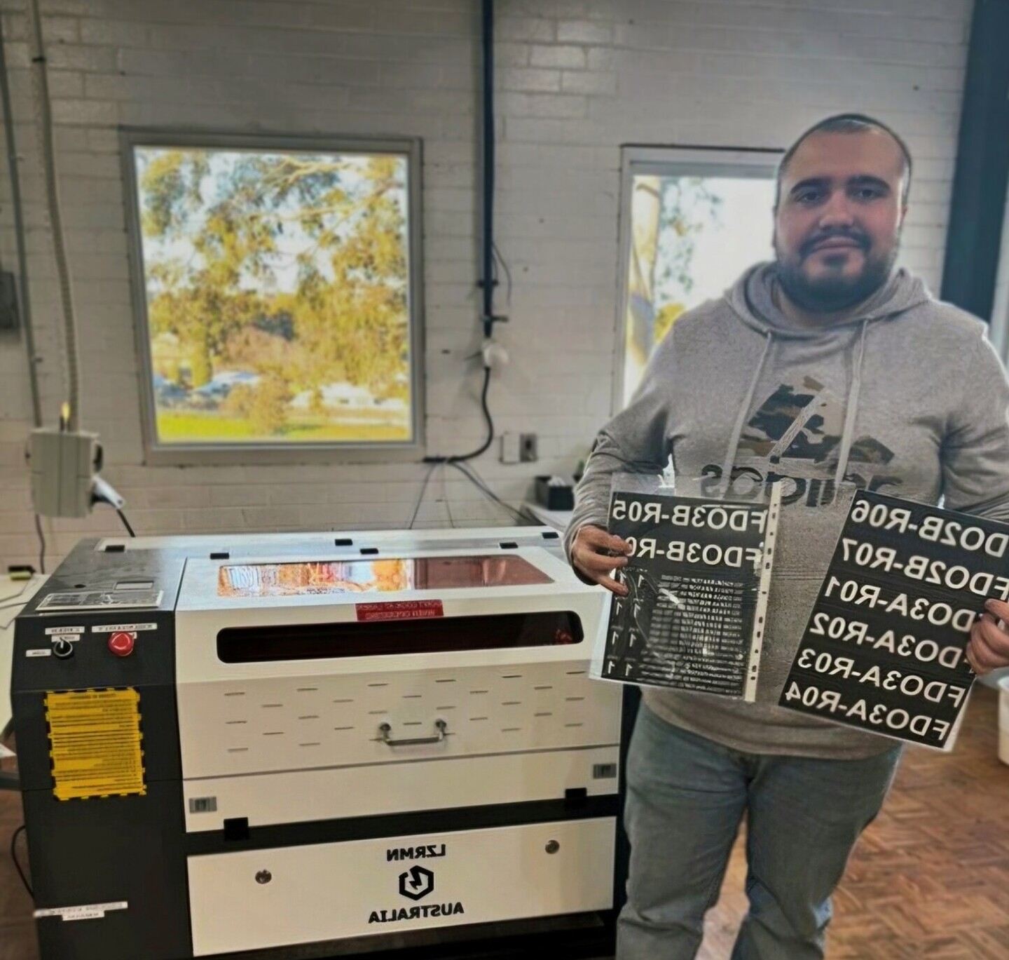 Andres is standing beside a laser cutting and engraving machine, showing off some recently made product.