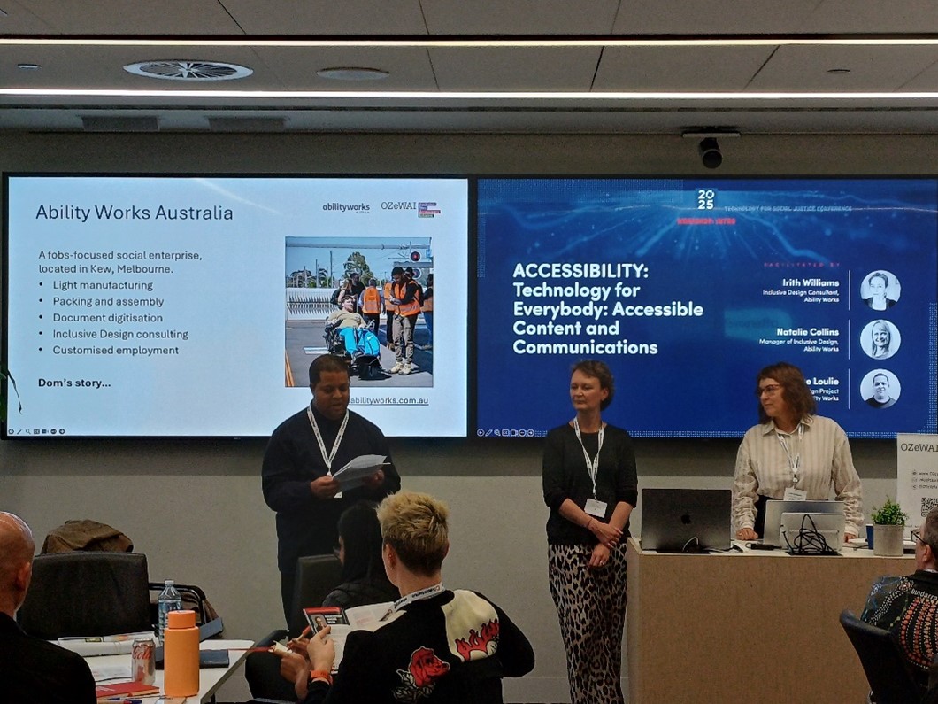 Three people (1 male and 2 women) are standing in front of seated people. The male is reading from a paper, with a screen behind them with the words, ‘Ability Works Australia’, and the title on the second screen, ‘Accessibility: Technology for everybody: Accessible Content and Communications’.