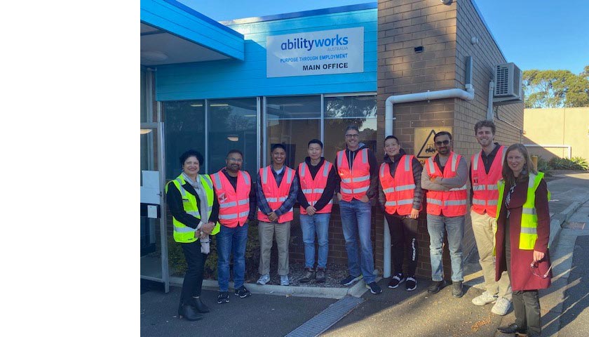 A group of seven Transurban team members stand outside the Ability Works office with AWA staff, Anu and Alana. Everyone is smiling and wearing bright hi-vis vests.