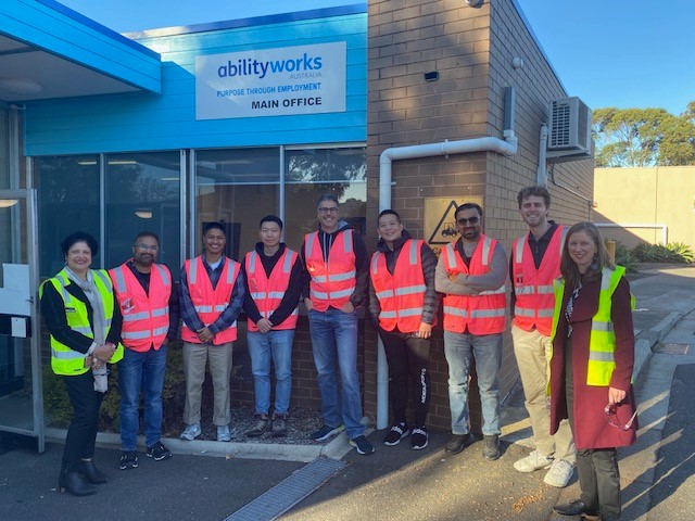 A group of seven Transurban team members stand outside the Ability Works office with AWA staff, Anu and Alana. Everyone is smiling and wearing bright hi-vis vests.