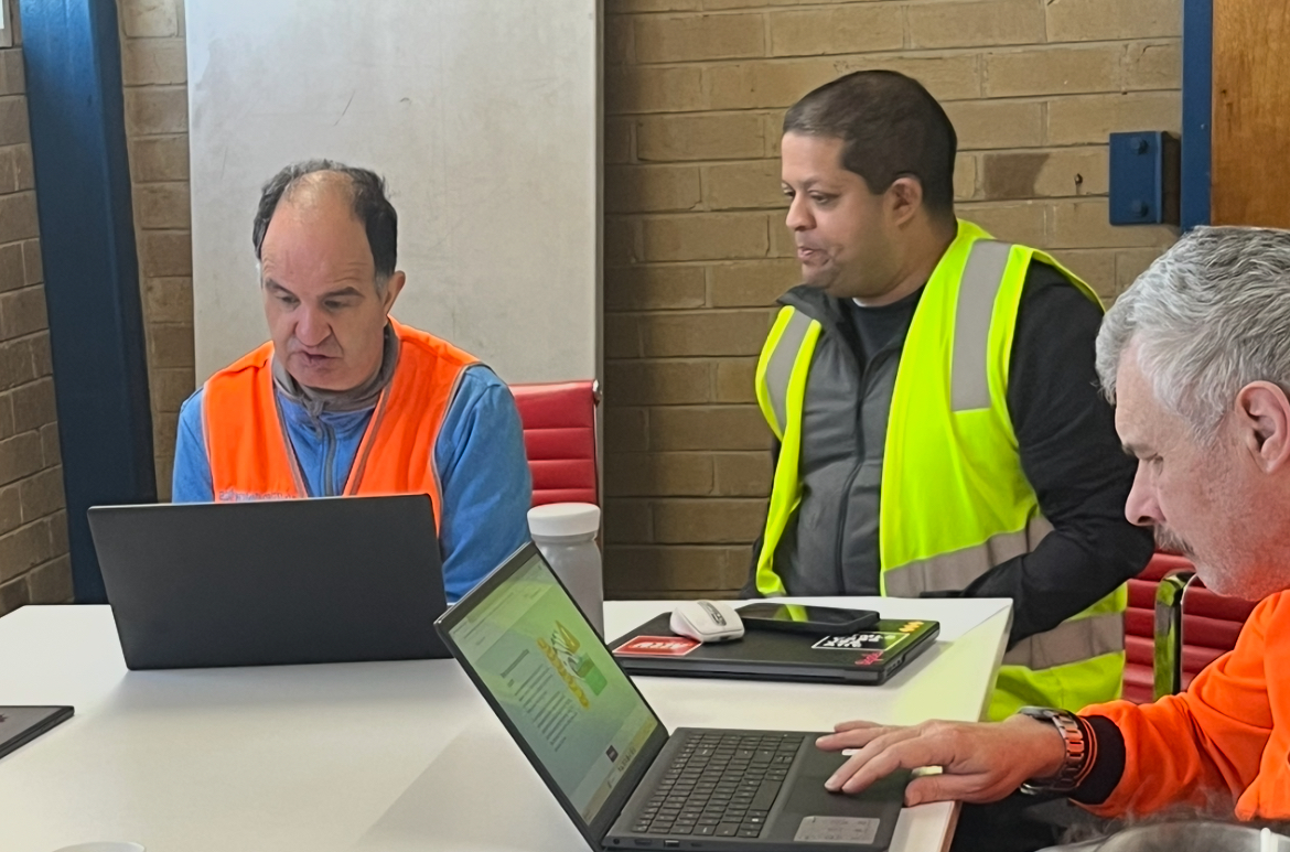 A man in a yellow high visibility vest looks at a screen with a man in an orange high visibility vest. Another man in the foreground uses the trackpad on a laptop.